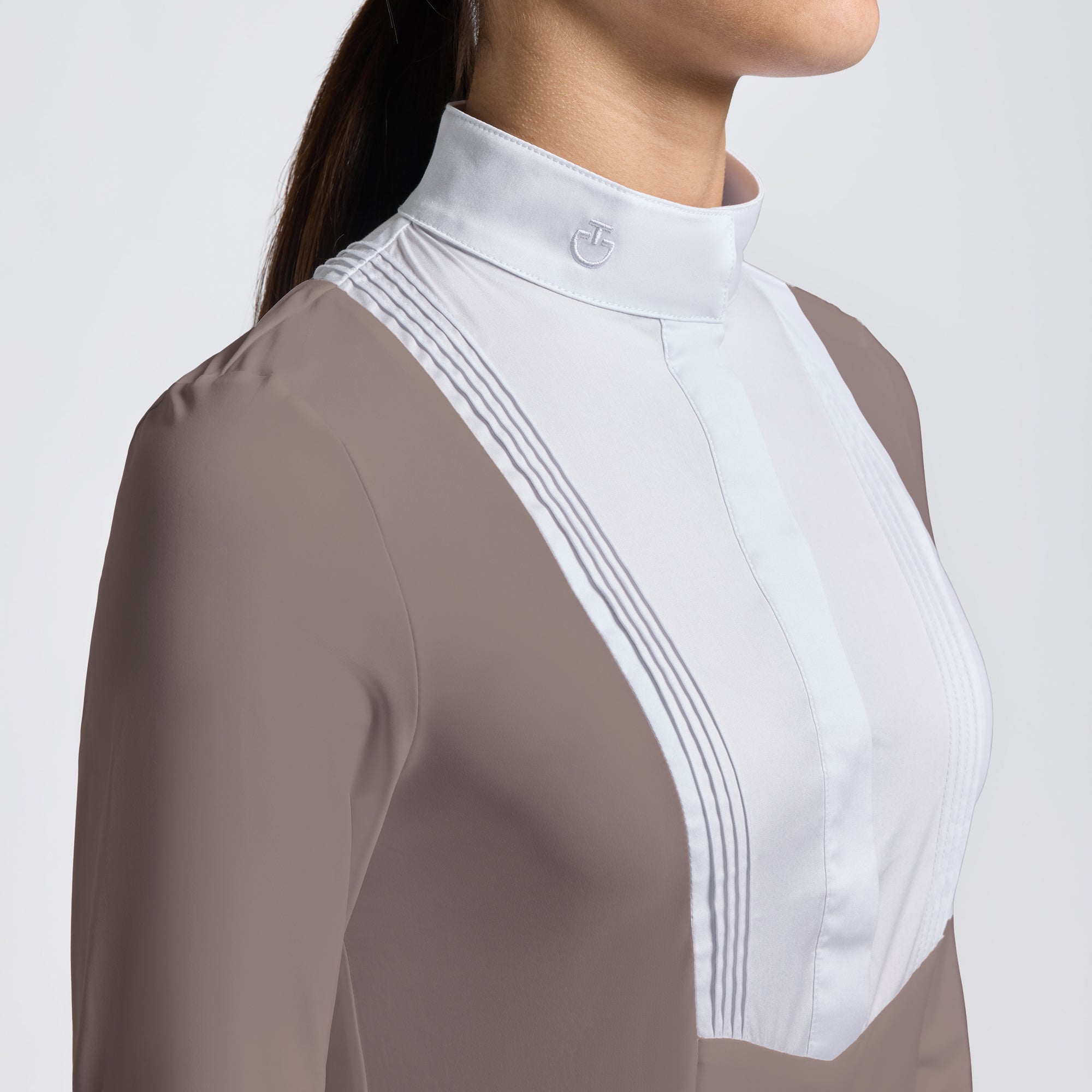 Close-up of a woman wearing a fitted long-sleeve equestrian-style top with a high mock neck. The garment is taupe brown with a contrasting white yoke panel featuring subtle vertical pleats and a small embroidered logo on the collar.