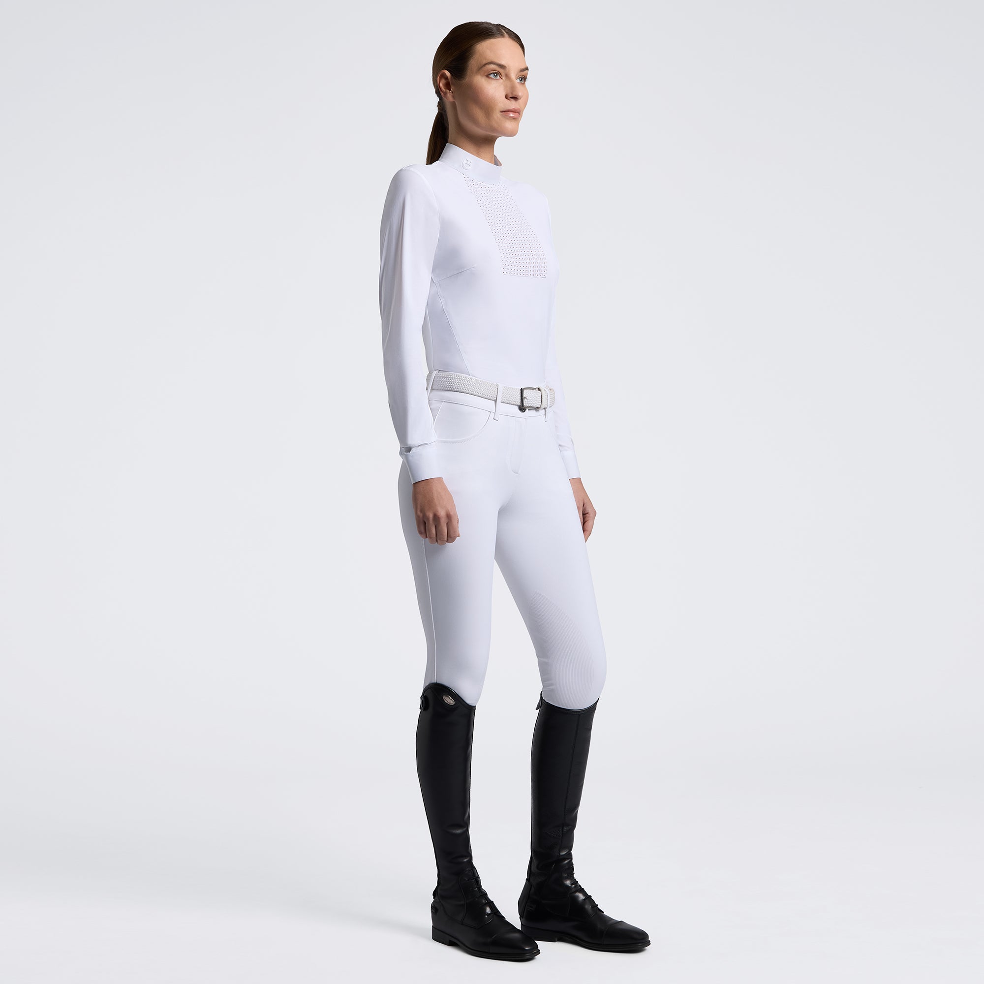A woman modeling a white long-sleeve equestrian-style top and white fitted pants with a silver belt, paired with black riding boots, against a clean light gray studio background.