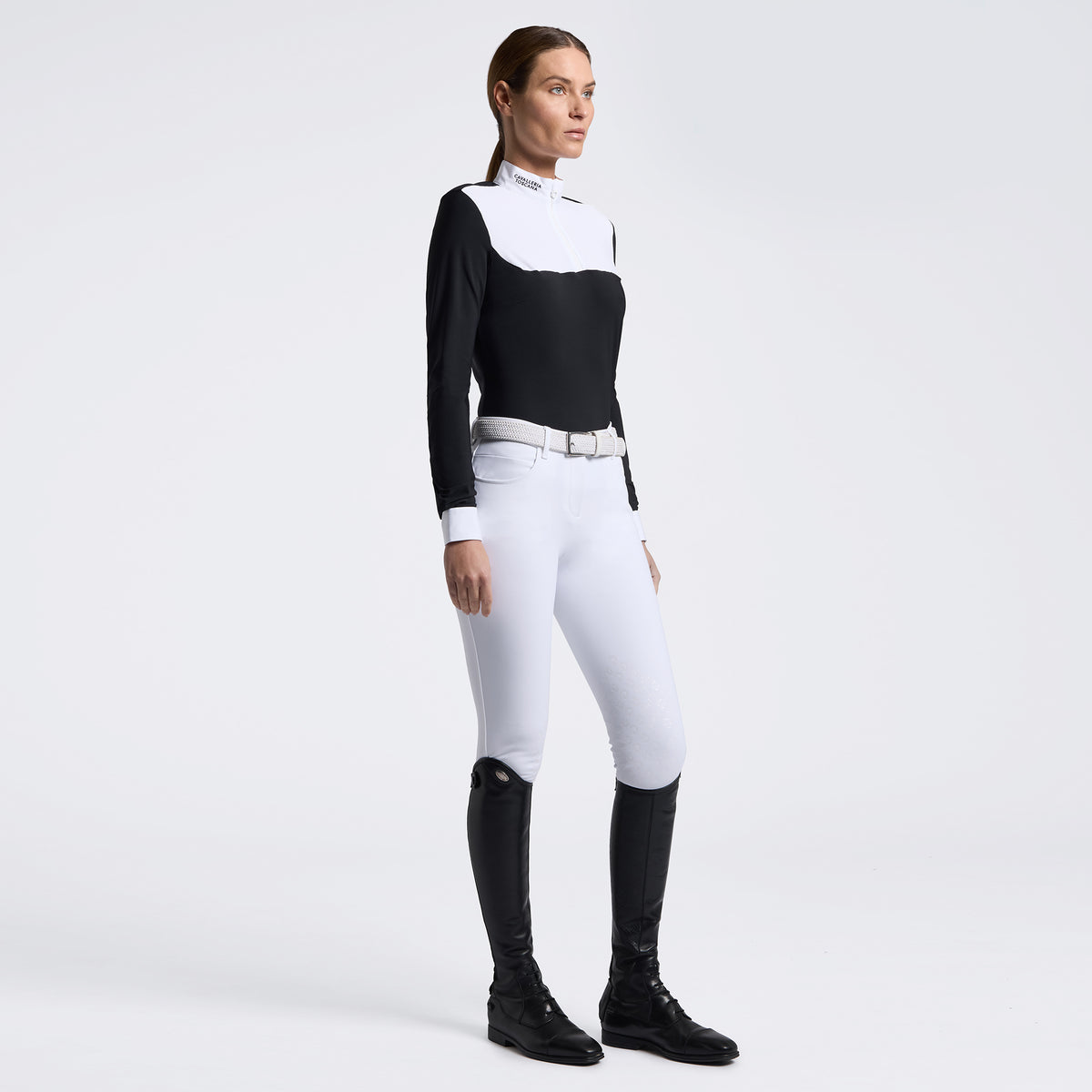 A woman modeling a black and white long-sleeve equestrian-style top with white riding breeches, a silver belt, and black riding boots against a light gray studio background.