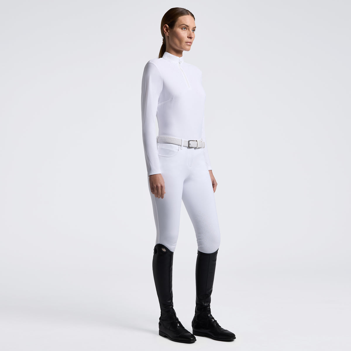 A woman modeling a white long-sleeve riding crop top and matching white breeches with a white belt, paired with black riding boots, against a light gray studio background.