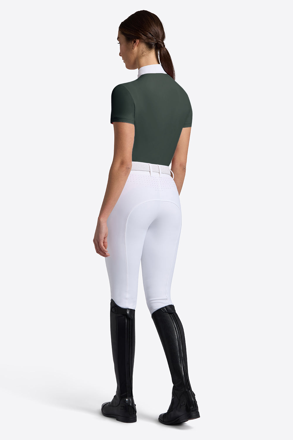 A woman shown from the back wearing fitted equestrian-style apparel: a dark green short-sleeve top, white riding breeches, and black tall riding boots against a clean light gray studio background.