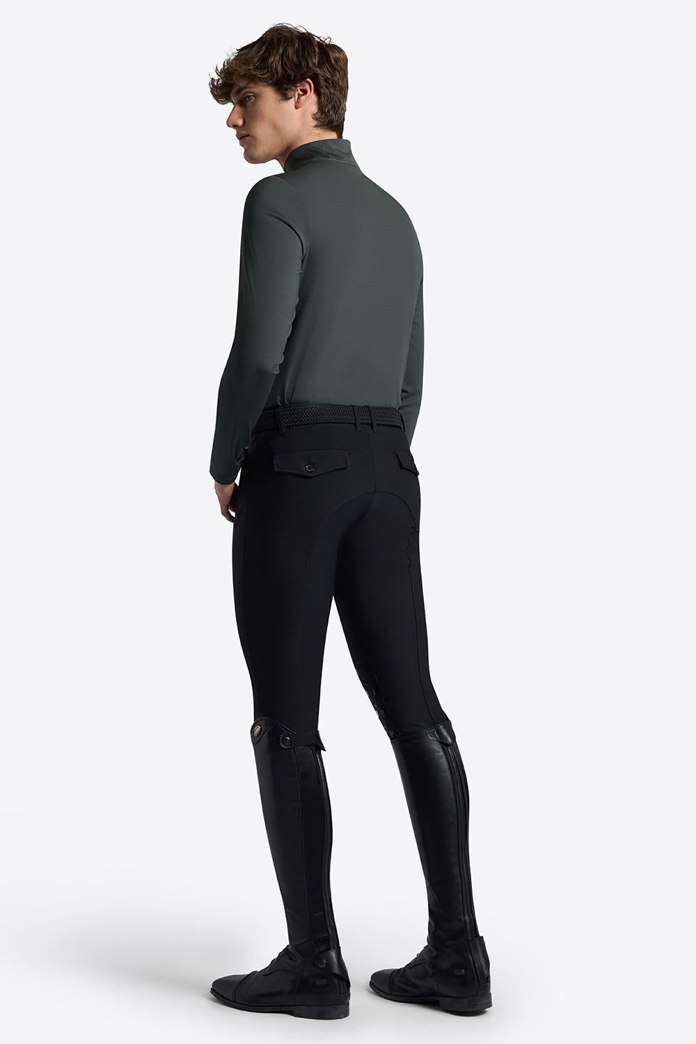 A man wearing a dark green mock-neck long-sleeve shirt and black riding breeches with black knee patches, shown from a rear three-quarter view against a light gray studio background.