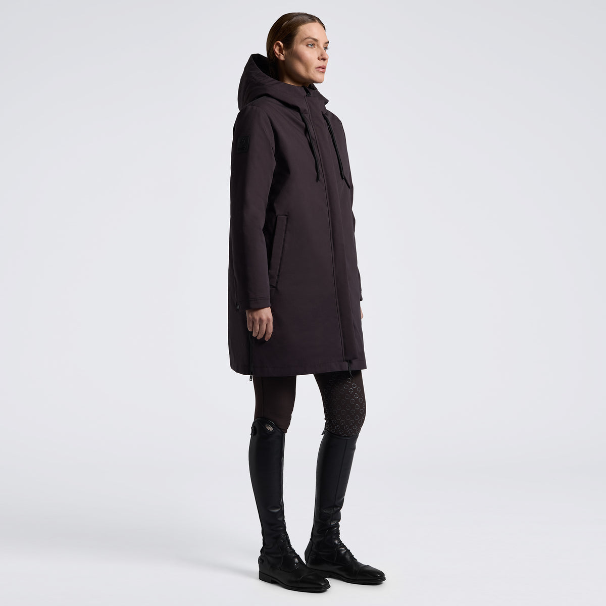 A model is shown in profile wearing a long, dark plum hooded parka jacket, styled with black patterned tights and black leather riding boots against a clean, light gray studio background.