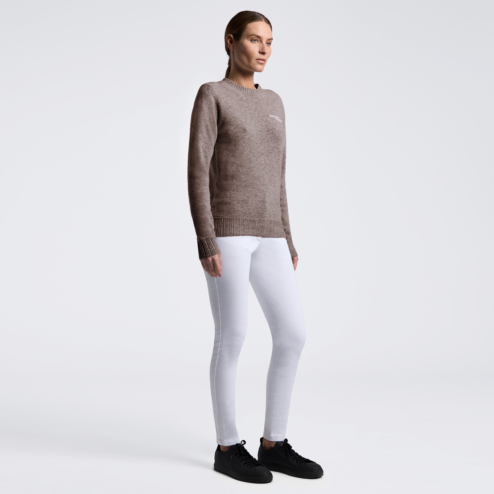A woman modeling a brown knit crewneck sweater with white leggings and black sneakers, standing against a light gray studio background.