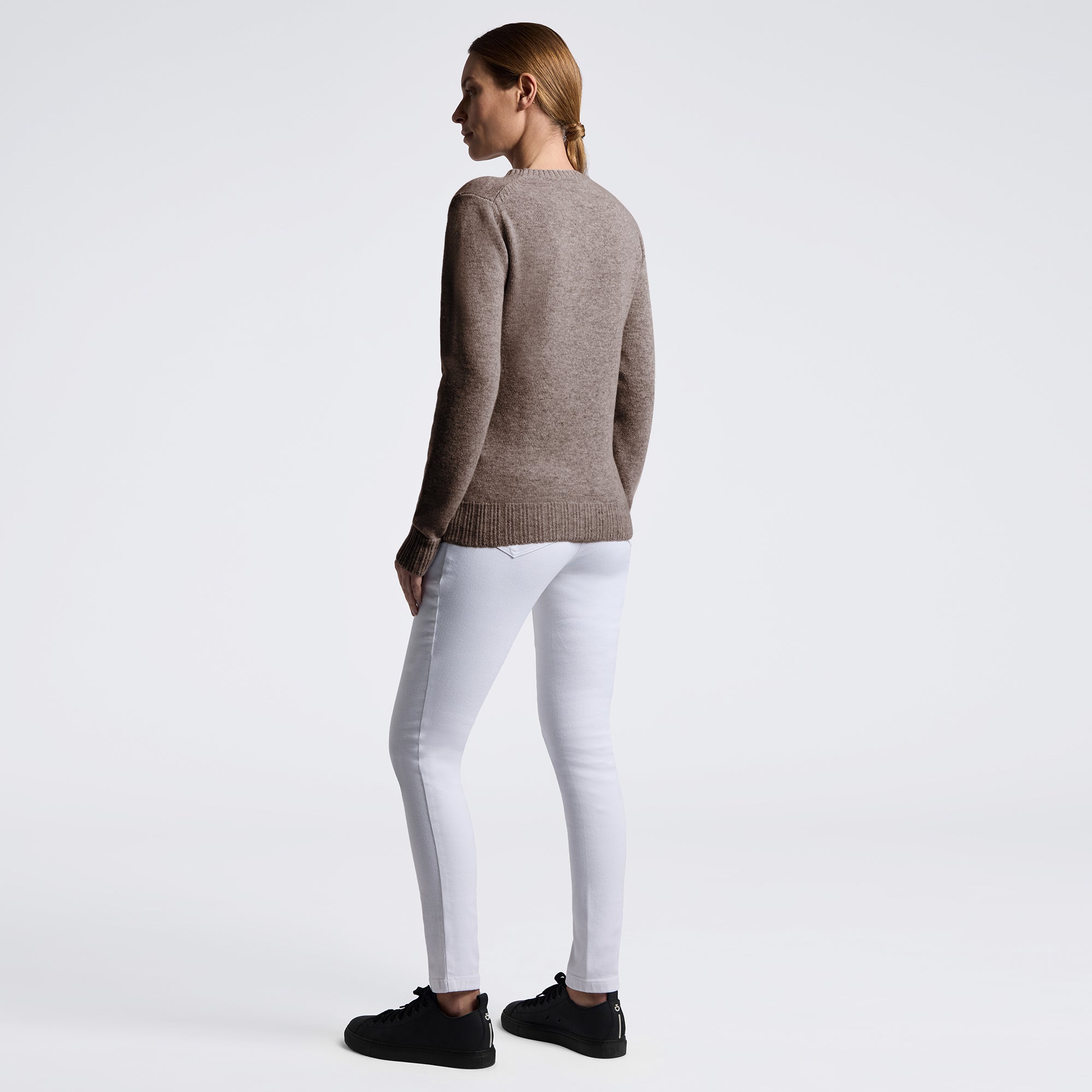 A woman shown from the back wearing a taupe knit sweater, white skinny jeans, and black sneakers against a plain white studio background.