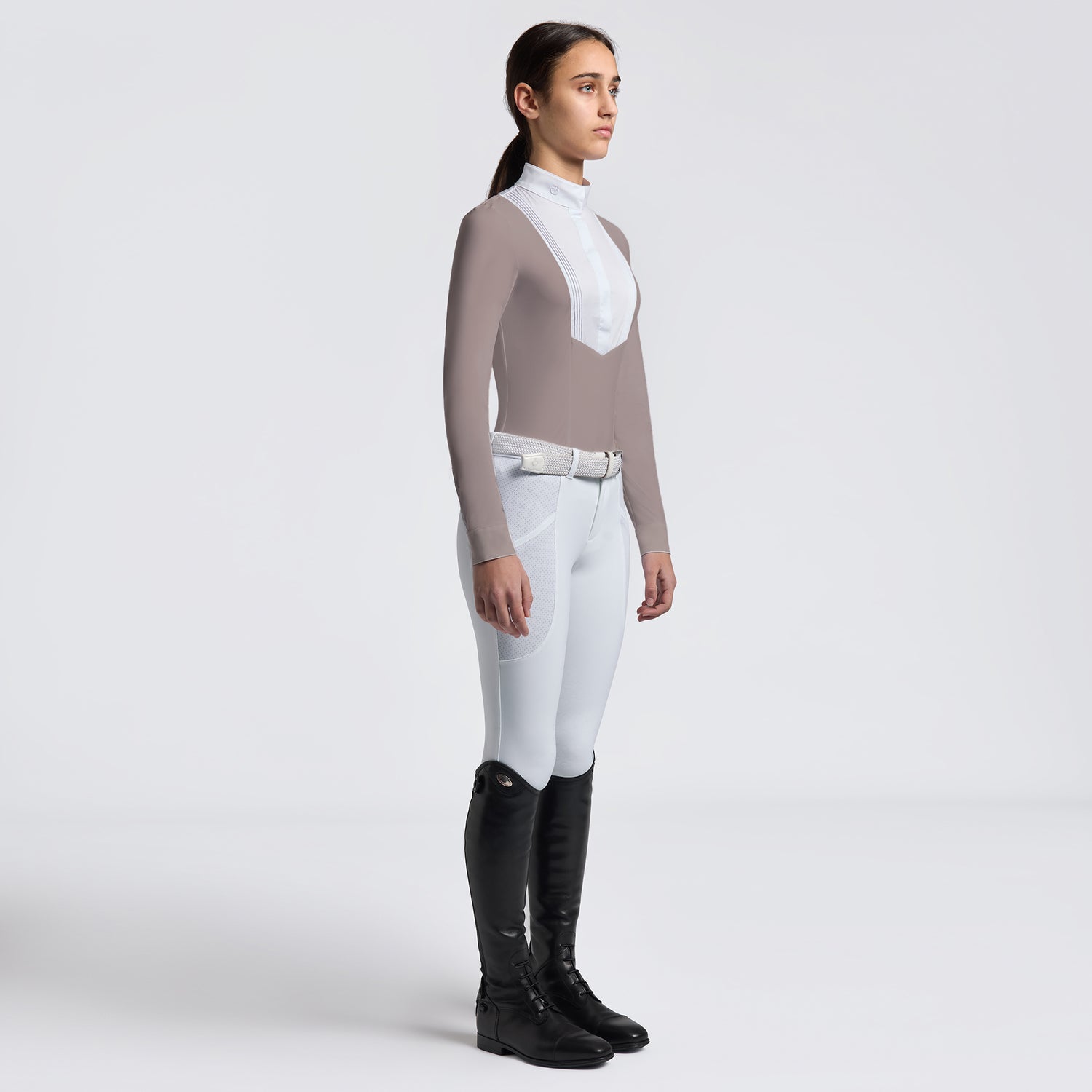 A woman stands in profile wearing a fitted long-sleeve equestrian-style top in taupe and white, white riding breeches, and tall black riding boots against a clean light gray studio background.