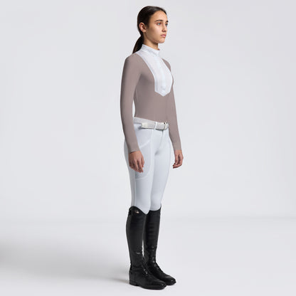 A woman stands in profile wearing a fitted long-sleeve equestrian-style top in taupe and white, white riding breeches, and tall black riding boots against a clean light gray studio background.