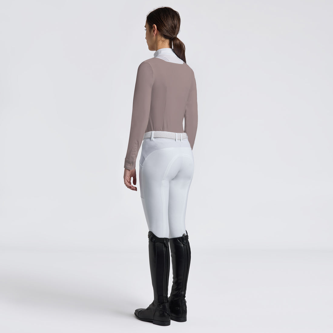 A woman shown from the back wearing a fitted taupe long-sleeve top, white riding breeches, and black equestrian boots against a clean light gray studio background.