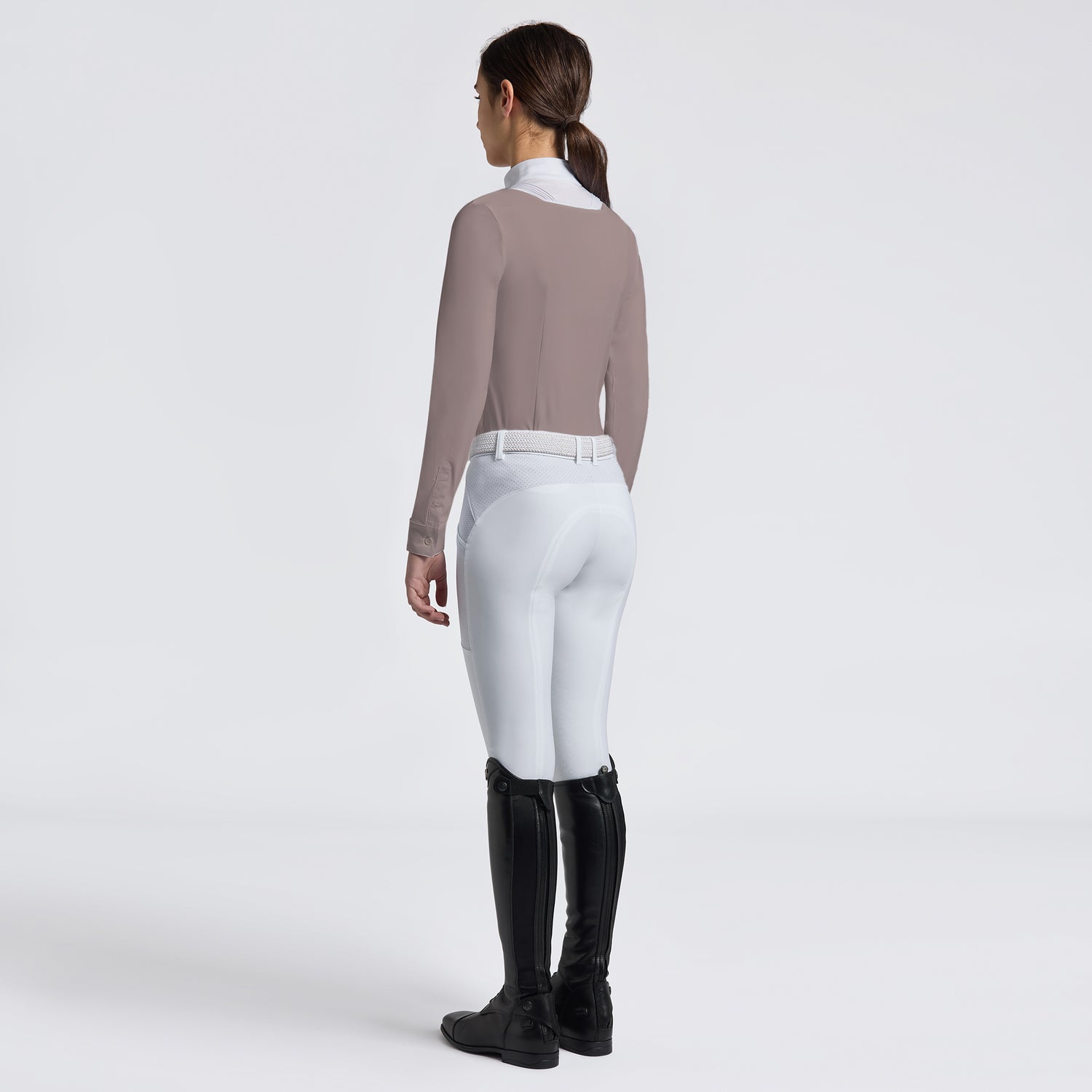 A woman shown from the back wearing a fitted taupe long-sleeve top, white riding breeches, and black equestrian boots against a clean light gray studio background.