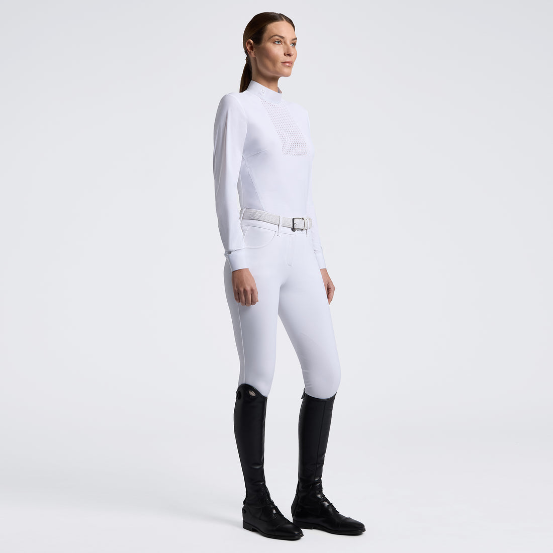 A woman modeling a white long-sleeve equestrian-style top and white fitted pants with a silver belt, paired with black riding boots, against a clean light gray studio background.