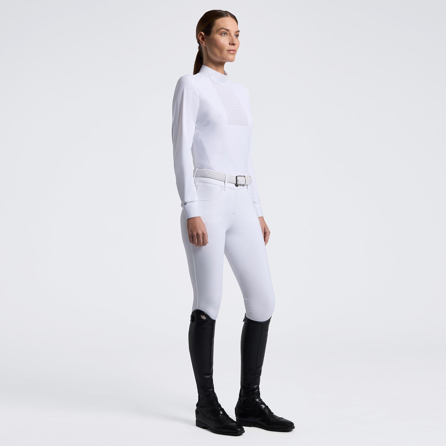 A woman modeling a white long-sleeve equestrian-style top and white fitted pants with a silver belt, paired with black riding boots, against a clean light gray studio background.
