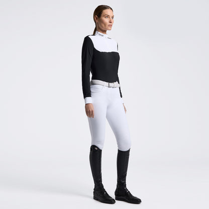 A woman modeling a black and white long-sleeve equestrian-style top with white riding breeches, a silver belt, and black riding boots against a light gray studio background.