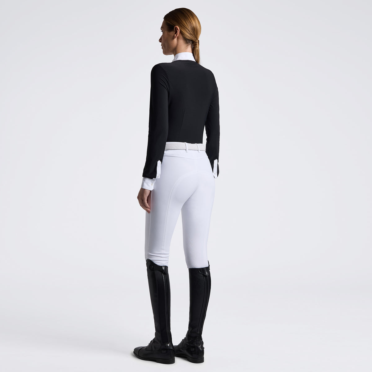 A woman shown from the back wearing a fitted black long-sleeve top, white riding breeches with a white belt, and black riding boots against a clean white studio background.