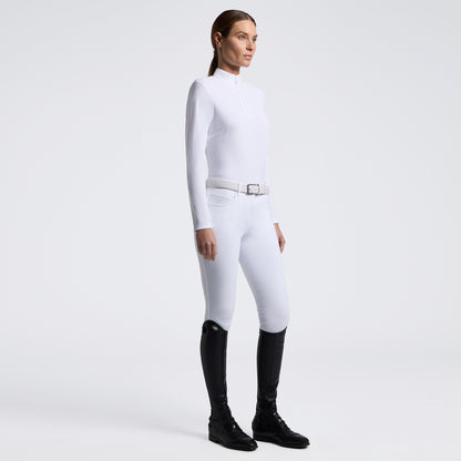 A woman modeling a white long-sleeve riding crop top and matching white breeches with a white belt, paired with black riding boots, against a light gray studio background.