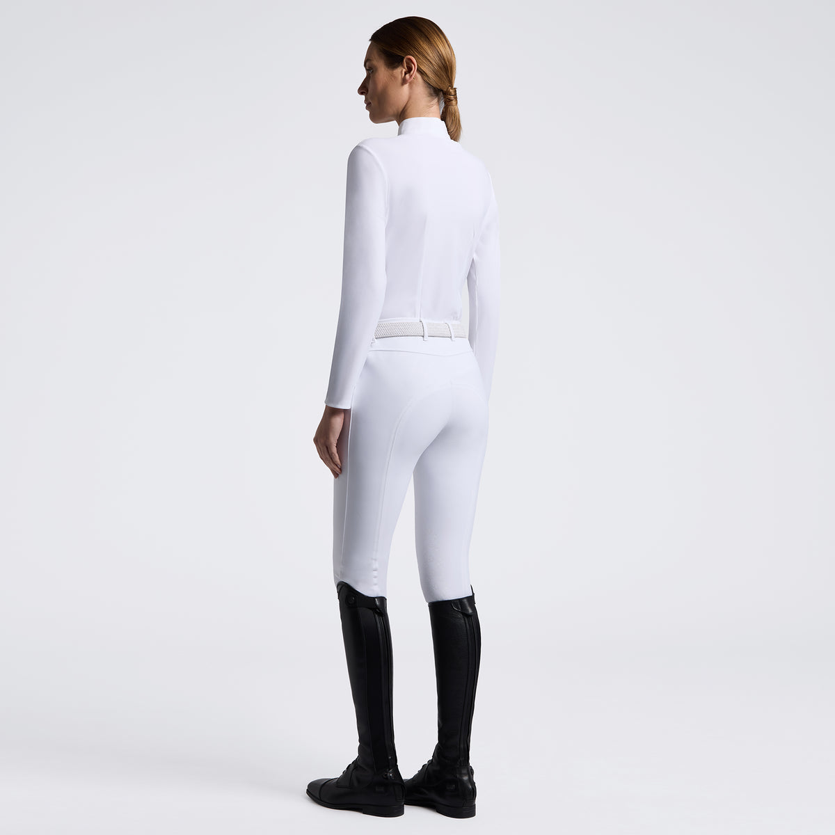 A woman shown from the back wearing a fitted white long-sleeve riding-style top and white breeches with a thin white belt, paired with black riding boots, against a clean light gray studio background.