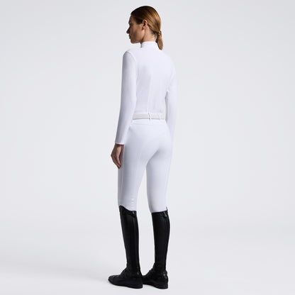 A woman shown from the back wearing a fitted white long-sleeve riding-style top and white breeches with a thin white belt, paired with black riding boots, against a clean light gray studio background.