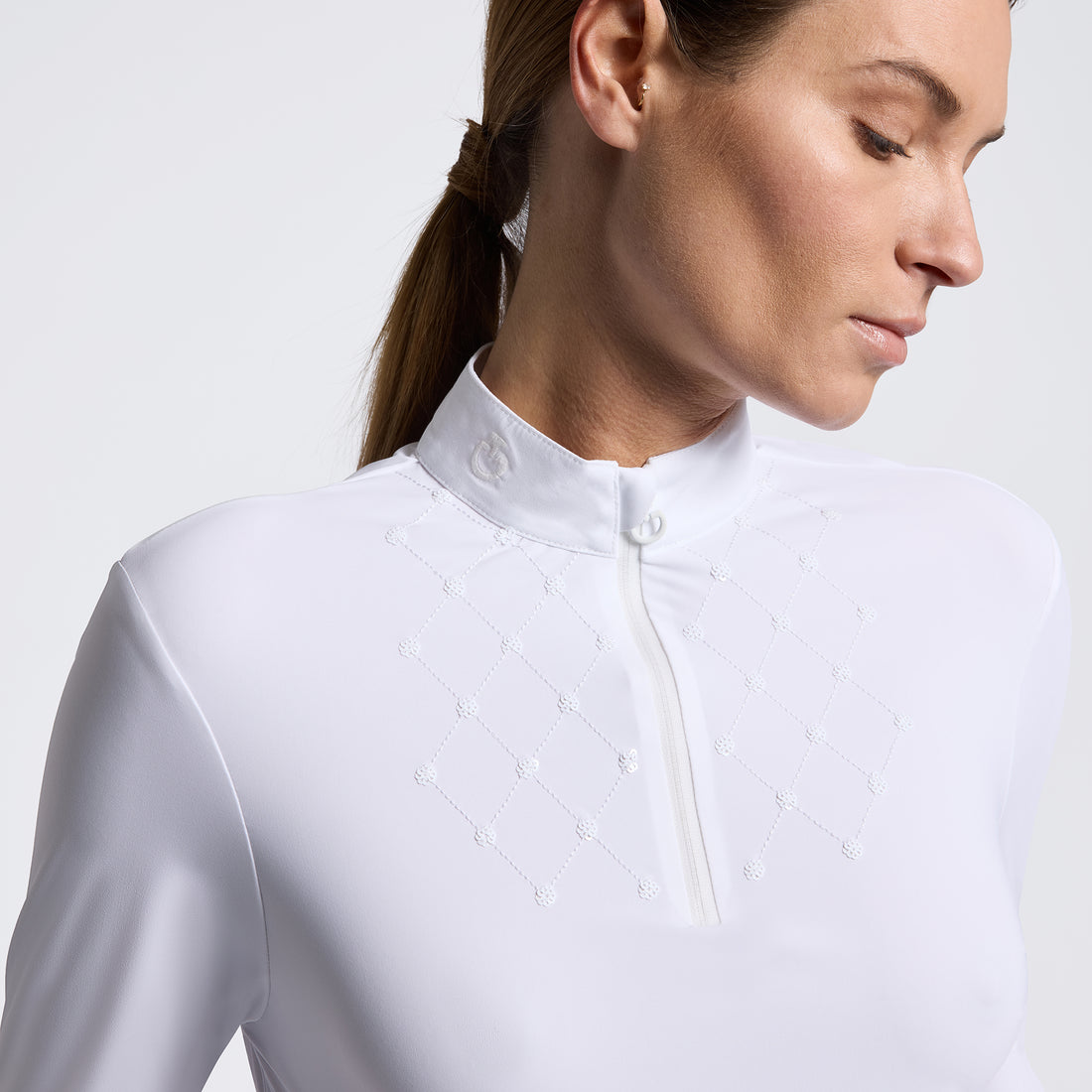 A close-up of a woman wearing a white equestrian-style half-zip mock neck jacket with delicate white embroidered geometric details on the chest, set against a clean light gray studio background.