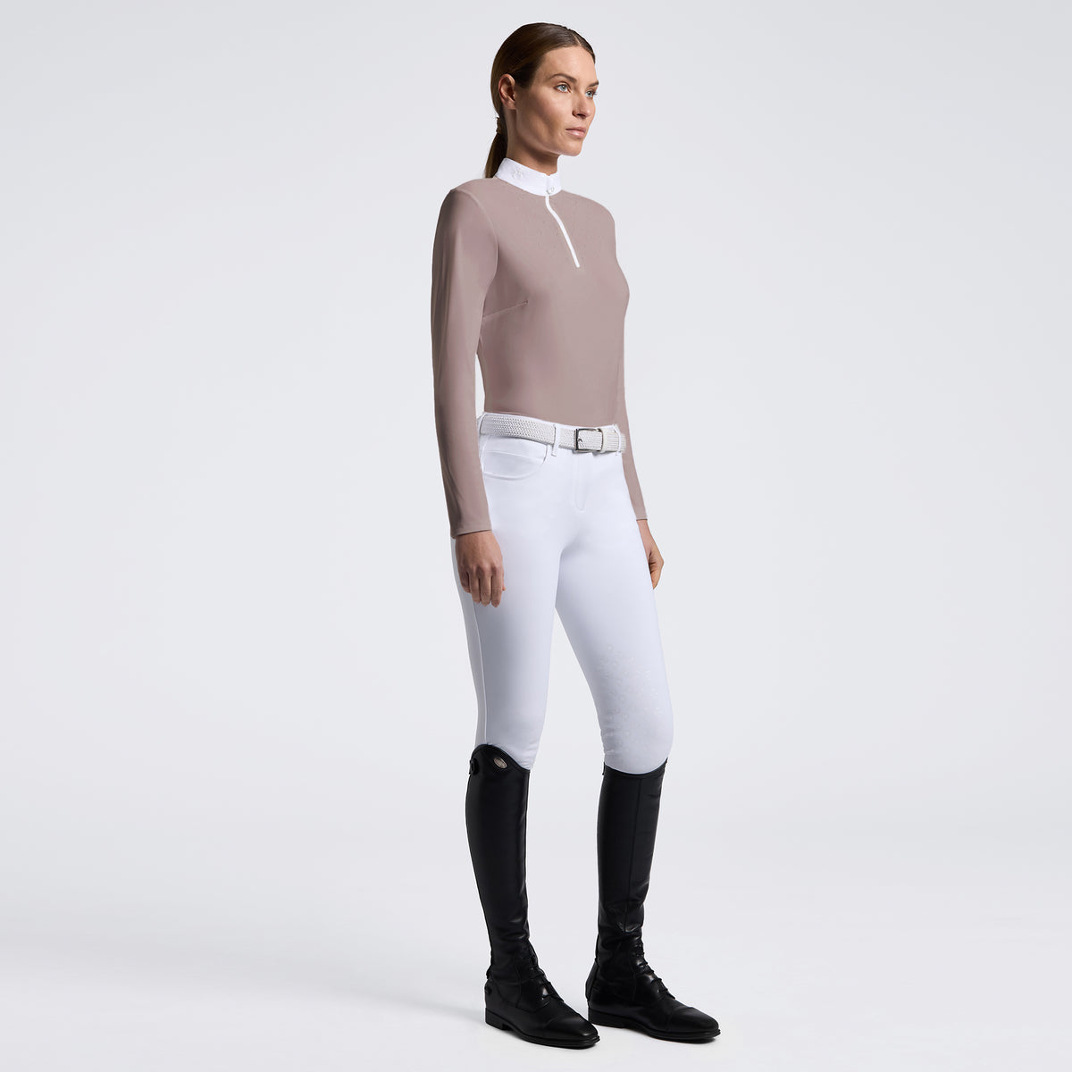 A woman stands in profile wearing a fitted long-sleeve quarter-zip top in taupe, white riding breeches with a silver belt, and black riding boots against a clean light gray studio background.