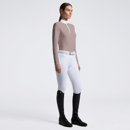 A woman stands in profile wearing a fitted long-sleeve quarter-zip top in taupe, white riding breeches with a silver belt, and black riding boots against a clean light gray studio background.