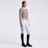 A woman stands in profile wearing a fitted long-sleeve quarter-zip top in taupe, white riding breeches with a silver belt, and black riding boots against a clean light gray studio background.