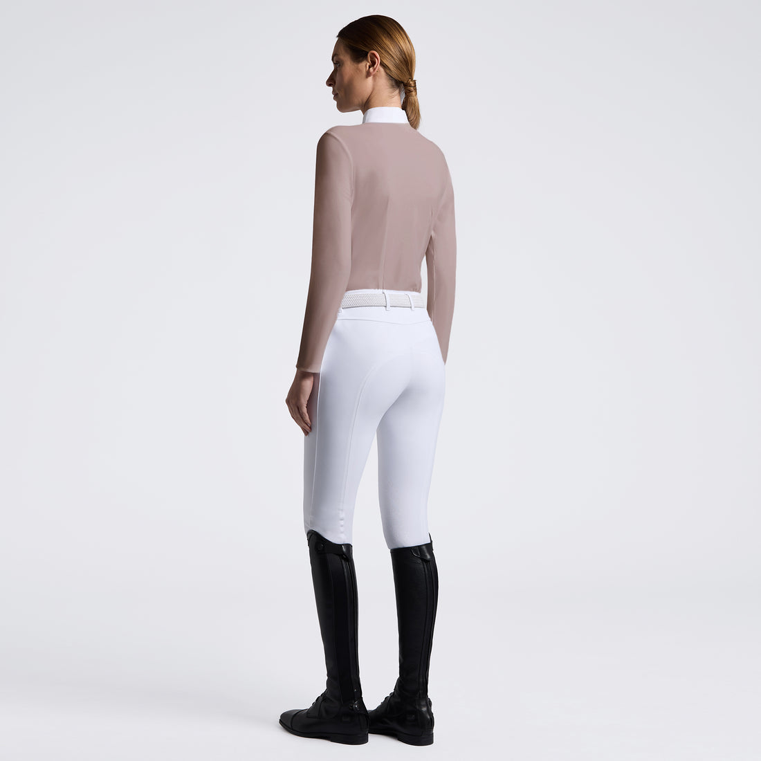 A woman shown from the back wearing white equestrian riding breeches, a fitted taupe long-sleeve top, and black riding boots against a clean light gray studio background.