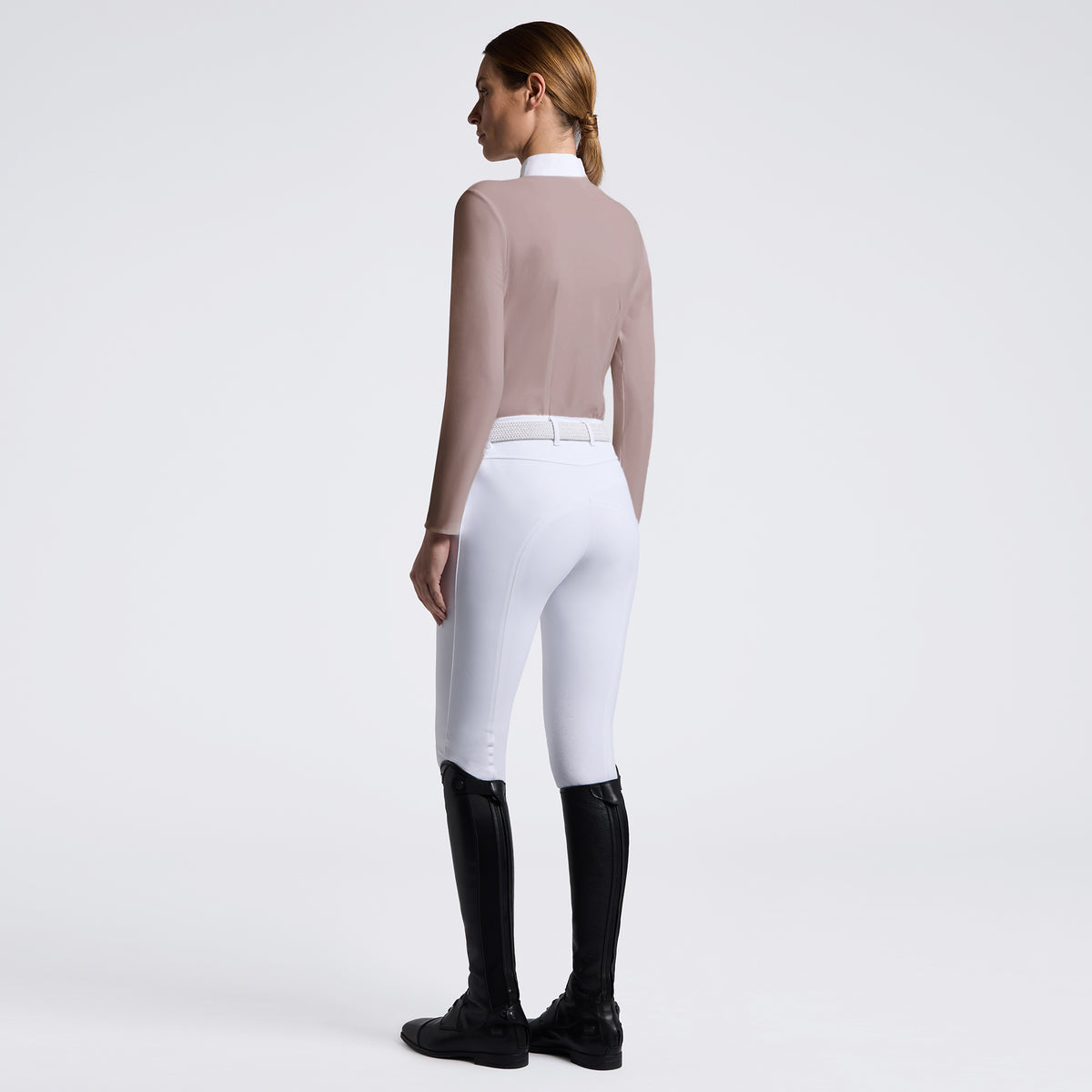 A woman shown from the back wearing white equestrian riding breeches, a fitted taupe long-sleeve top, and black riding boots against a clean light gray studio background.