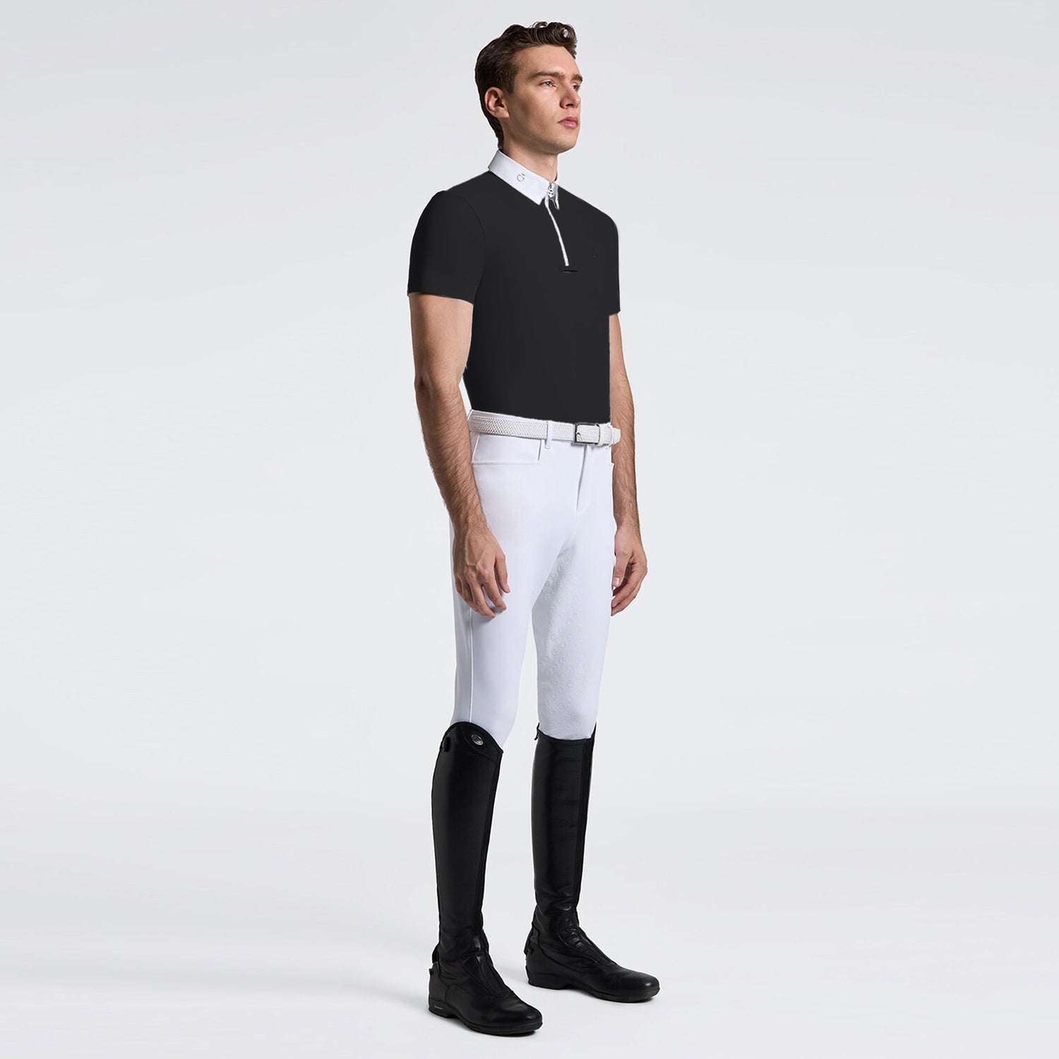 A male model stands in profile wearing a black short-sleeve polo shirt with a white collar and half-zip, white riding breeches, a white belt, and black riding boots against a light gray studio background.