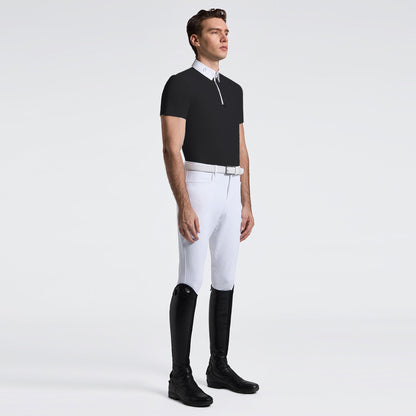 A male model stands in profile wearing a black short-sleeve polo shirt with a white collar and half-zip, white riding breeches, a white belt, and black riding boots against a light gray studio background.