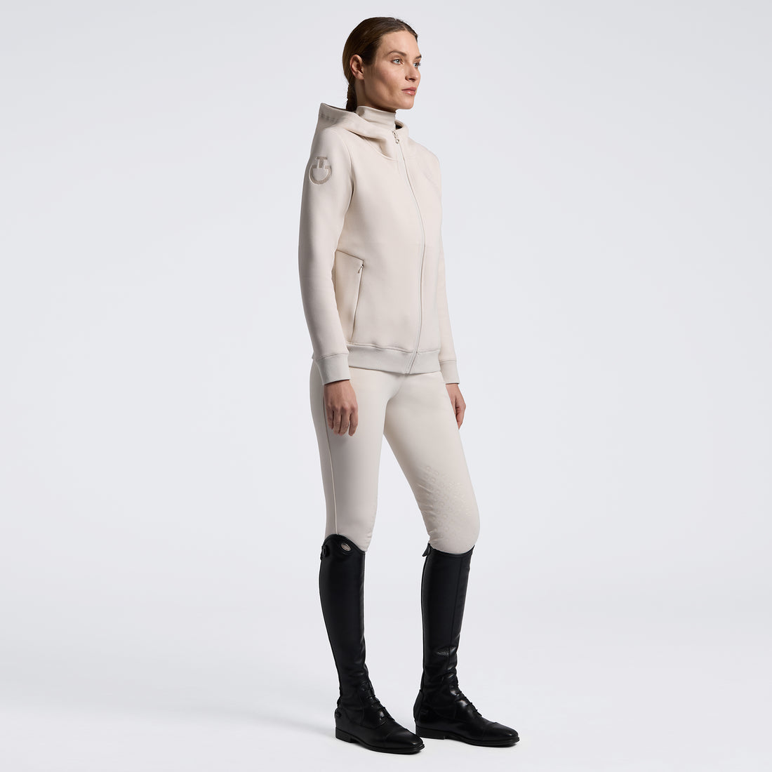 A model stands in profile wearing a light warm off-white equestrian riding jacket and matching breeches, paired with black riding boots, against a seamless light gray studio background.