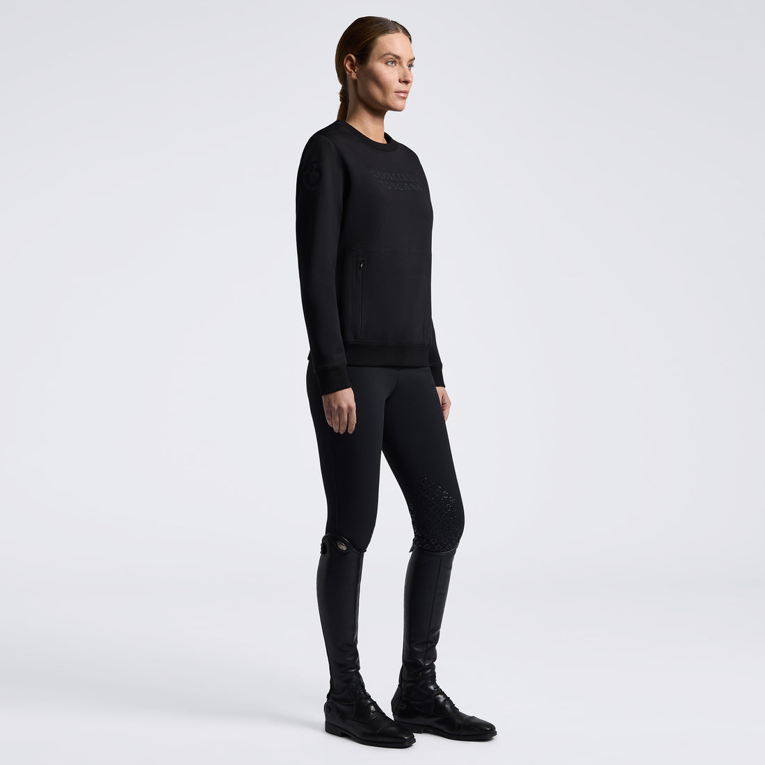 A woman stands in profile wearing a black long-sleeve crewneck sweatshirt, black fitted leggings with subtle textured details on the legs, and black boots against a clean light gray studio background.