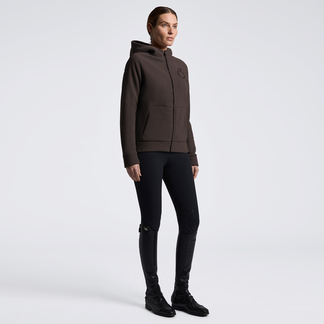 A model wearing a dark brown zip-up hoodie with a small tonal logo on the left chest, paired with black skinny pants and black boots, standing against a light gray studio background.