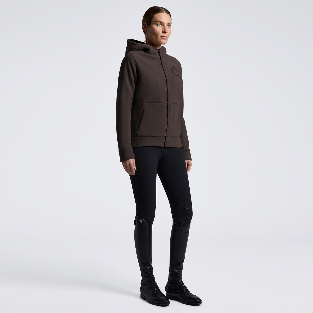 A model wearing a dark brown zip-up hoodie with a small tonal logo on the left chest, paired with black skinny pants and black boots, standing against a light gray studio background.