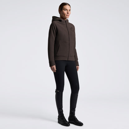 A model wearing a dark brown zip-up hoodie with a small tonal logo on the left chest, paired with black skinny pants and black boots, standing against a light gray studio background.