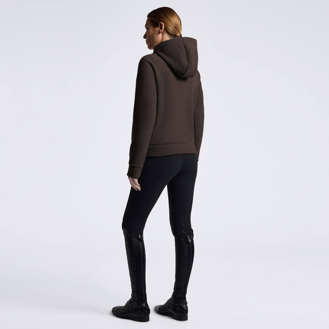 A woman shown from the back wearing a dark brown pullover hoodie, black fitted pants, and black knee-high boots against a clean light gray studio background.