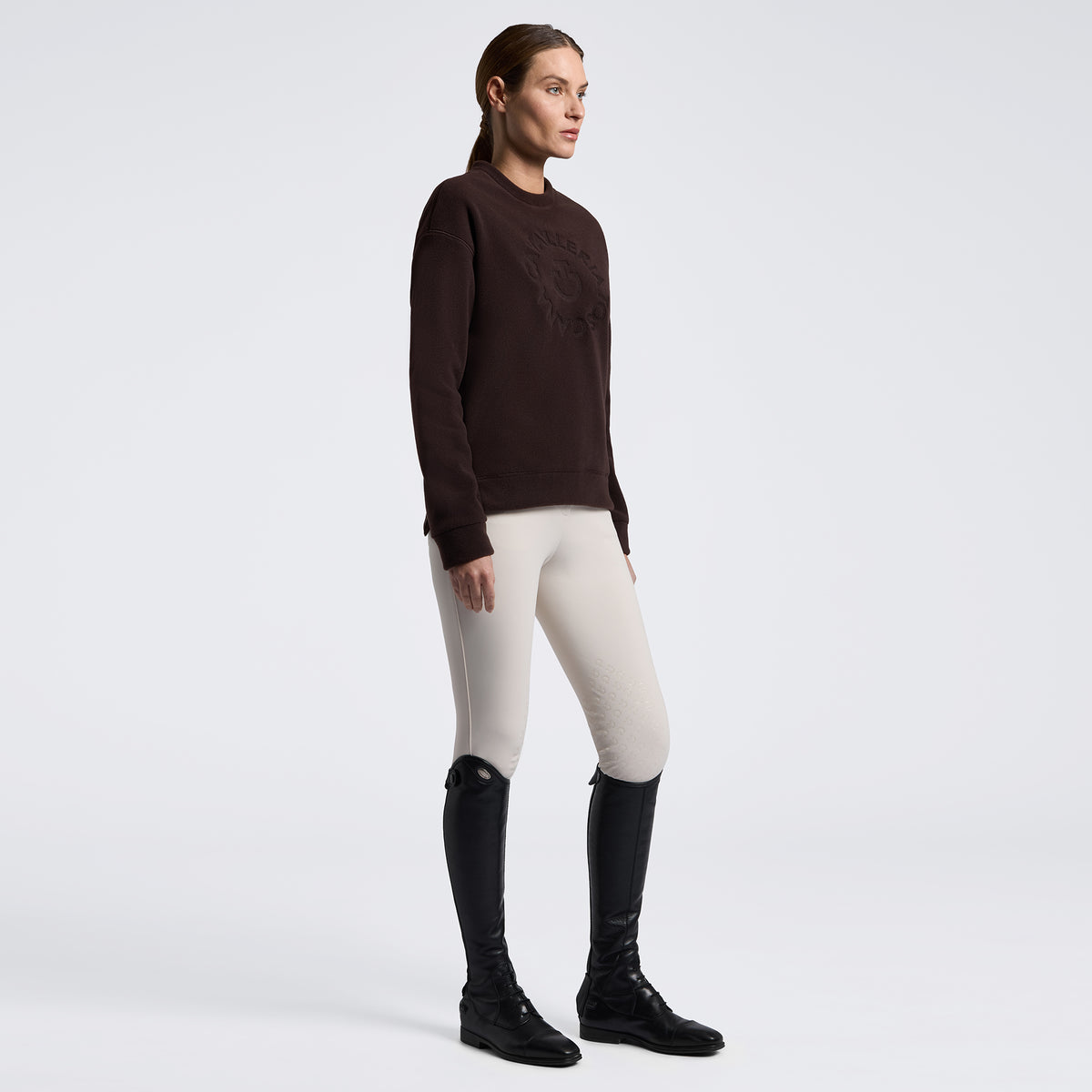 A woman stands in profile wearing a dark brown knit sweater with tonal embroidery, light beige riding breeches, and black riding boots against a clean white studio background.