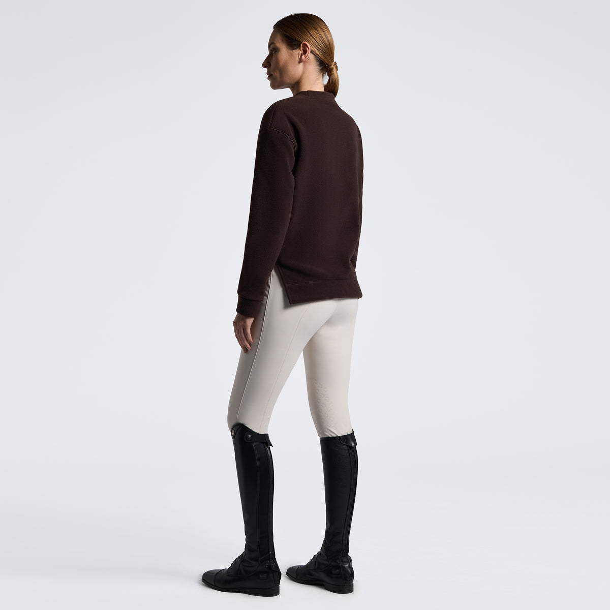 A woman shown from the back wearing a dark brown mock-neck long-sleeve top, light beige riding breeches, and black riding boots against a plain white studio background.