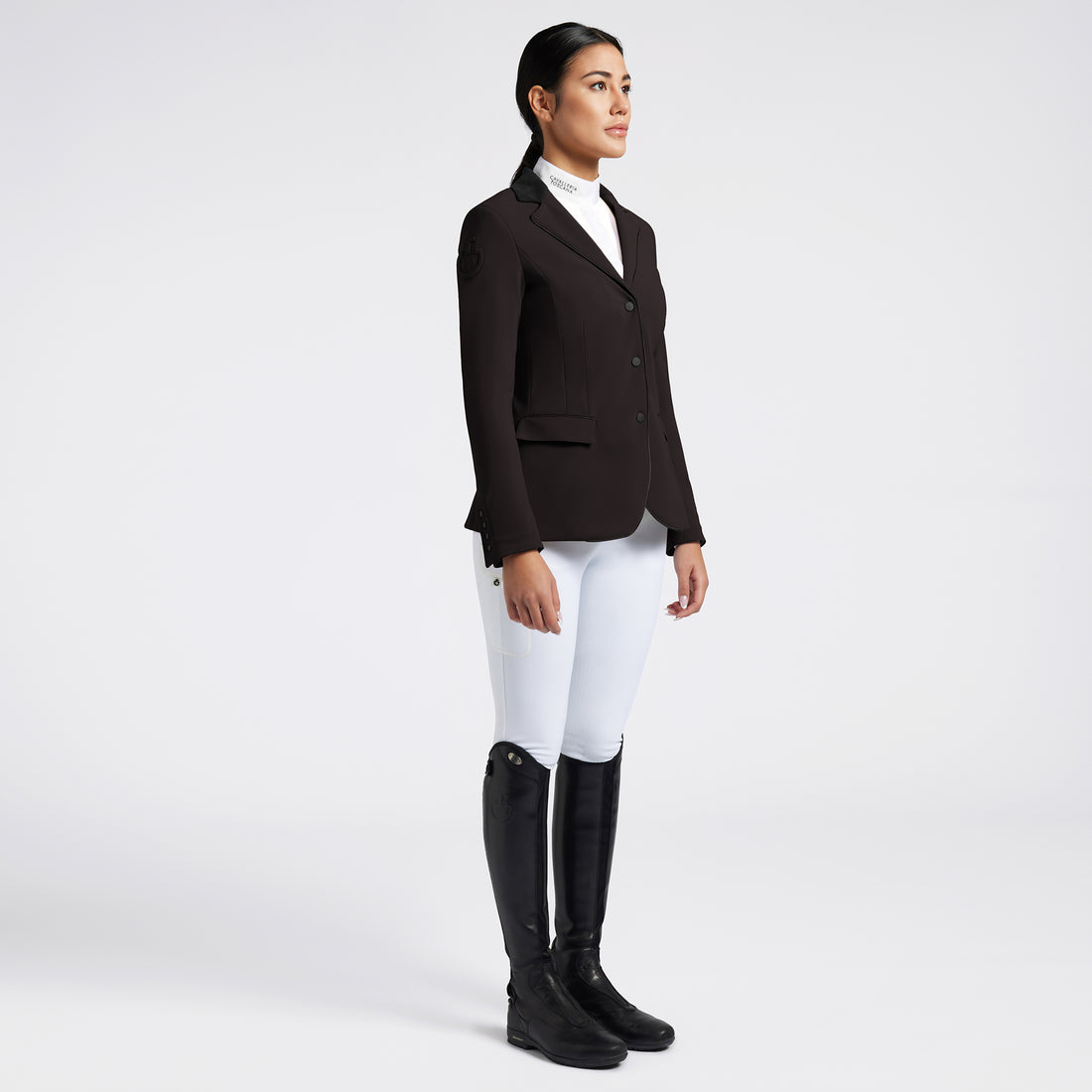 A woman stands in profile wearing a dark brown equestrian blazer, white riding breeches, and black riding boots against a light gray studio background.
