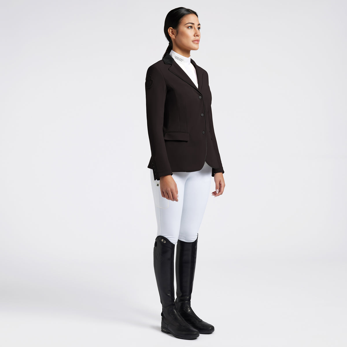 A woman stands in profile wearing a dark brown equestrian blazer, white riding breeches, and black riding boots against a light gray studio background.