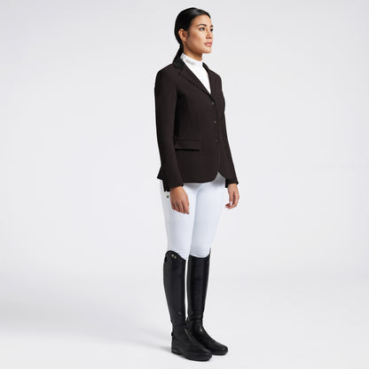 A woman stands in profile wearing a dark brown equestrian blazer, white riding breeches, and black riding boots against a light gray studio background.