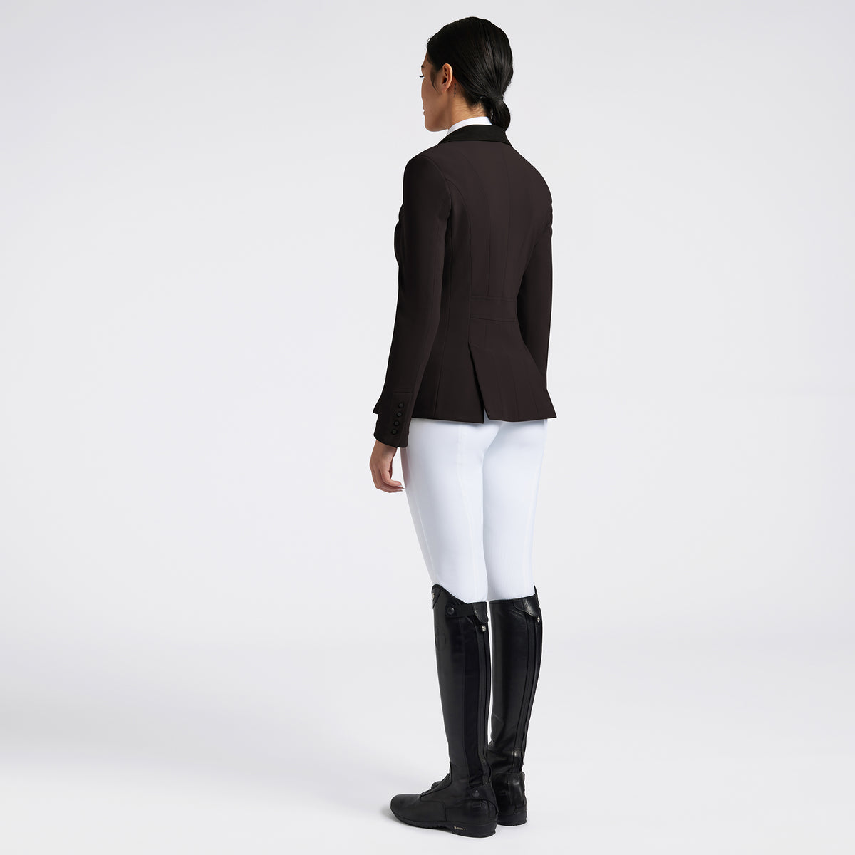 A woman shown from the back wearing a dark brown equestrian jacket, white breeches, and black riding boots against a light gray studio background.