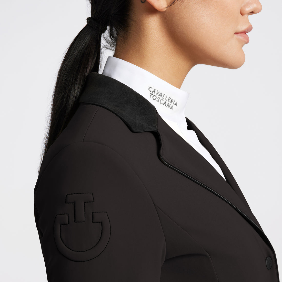 Close-up side profile of a woman wearing a brown  equestrian-style jacket with a black collar, a white collared shirt with &