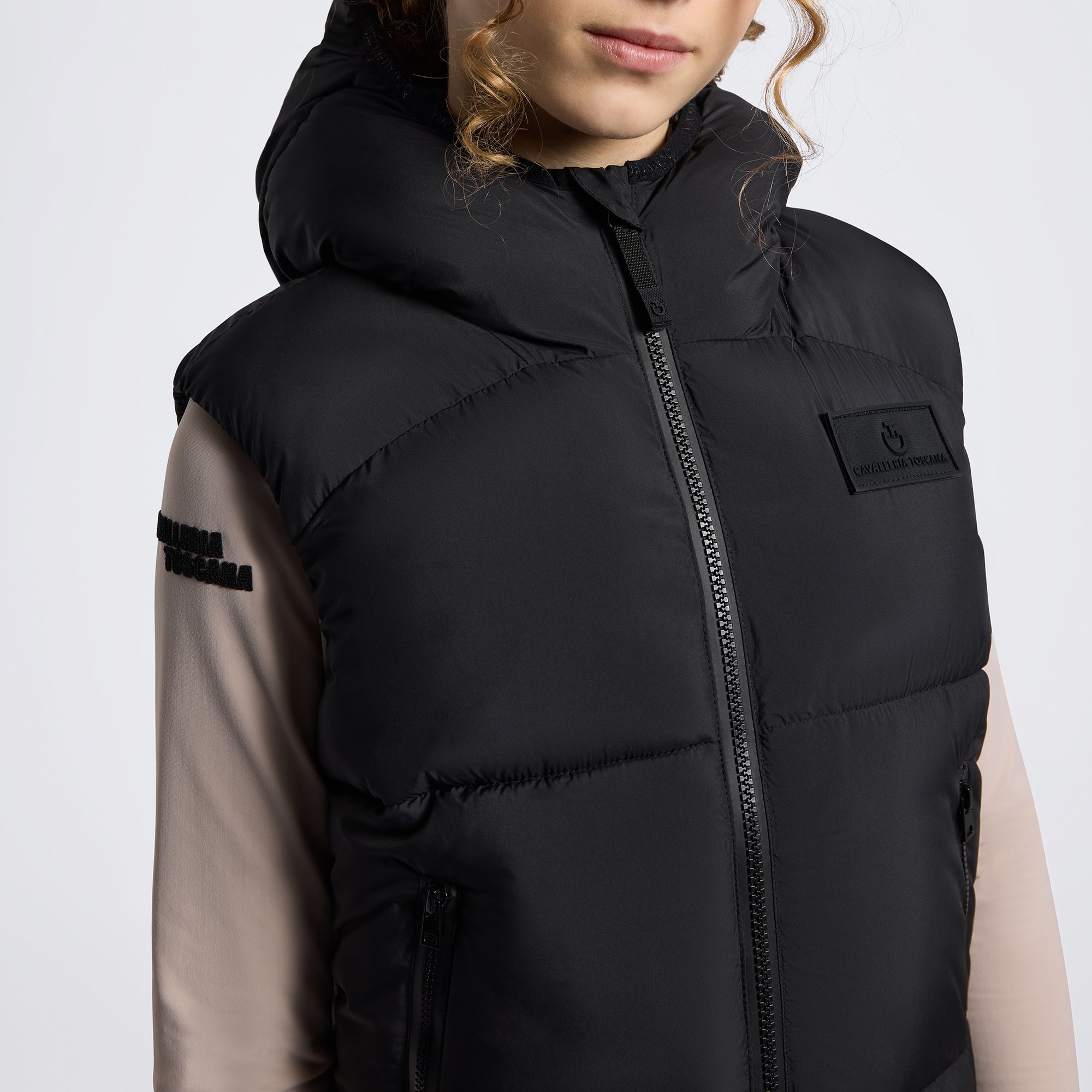 A close-up of a person wearing a black hooded puffer vest over a beige long-sleeve top. The vest features a front zipper, horizontal quilting, a small rectangular logo patch on the left chest, and black text on the right sleeve.