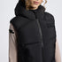 A close-up of a person wearing a black hooded puffer vest over a beige long-sleeve top. The vest features a front zipper, horizontal quilting, a small rectangular logo patch on the left chest, and black text on the right sleeve.