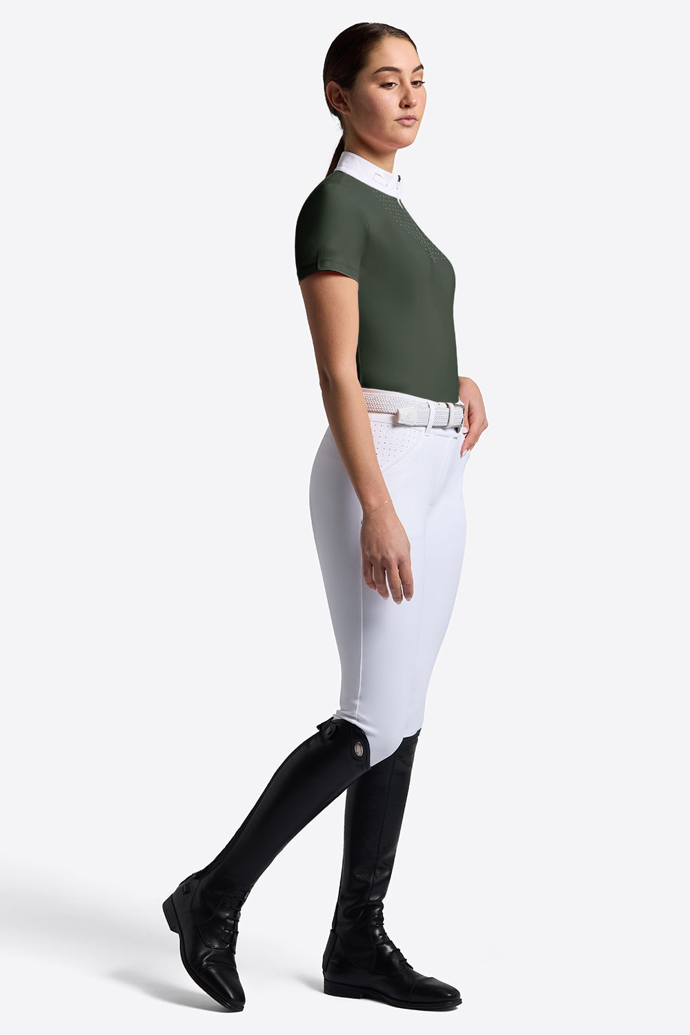 A woman modeling a dark green short-sleeve equestrian shirt with a white collar, paired with white riding breeches, a white belt, and black riding boots against a light gray studio background.