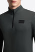 A close-up of a man wearing a dark green long-sleeve quarter-zip athletic top with a black rectangular logo patch on the chest.
