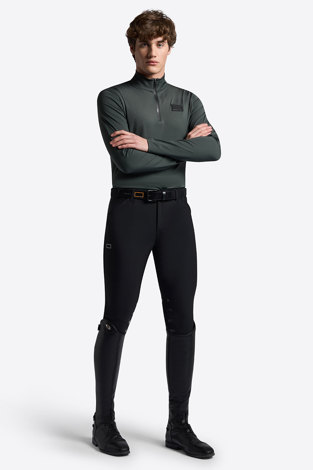A male model wearing a dark green long-sleeve quarter-zip performance top, black riding breeches, and black riding boots, standing with arms crossed against a light gray studio background.