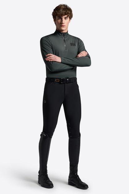 A male model wearing a dark green long-sleeve quarter-zip performance top, black riding breeches, and black riding boots, standing with arms crossed against a light gray studio background.