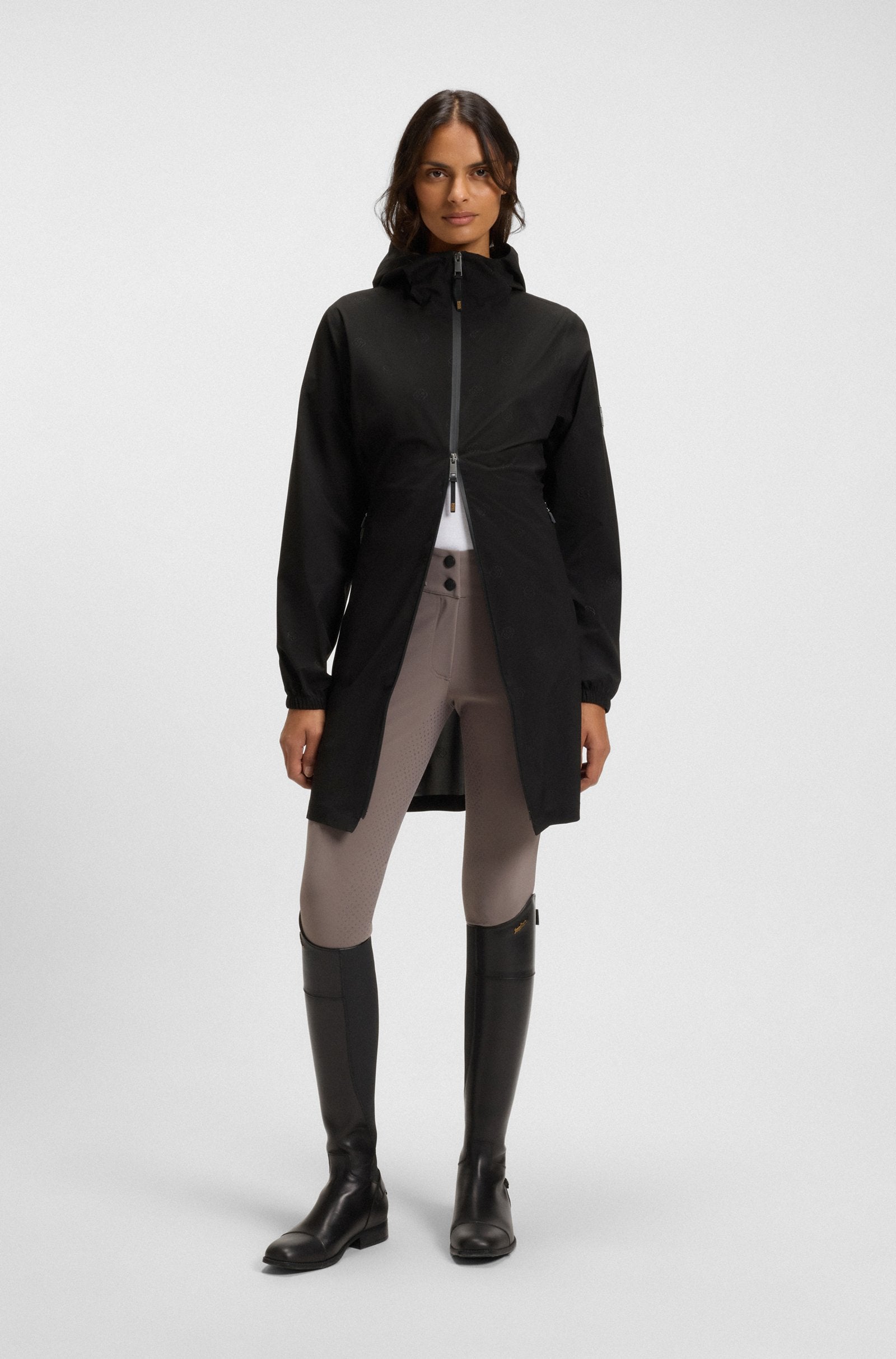 A model stands against a light gray studio background wearing a black hooded zip-front jacket with a gathered waist, light gray riding breeches, and tall black riding boots.