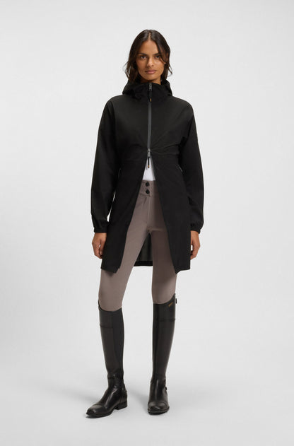 A model stands against a light gray studio background wearing a black hooded zip-front jacket with a gathered waist, light gray riding breeches, and tall black riding boots.