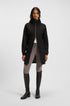 A model stands against a light gray studio background wearing a black hooded zip-front jacket with a gathered waist, light gray riding breeches, and tall black riding boots.
